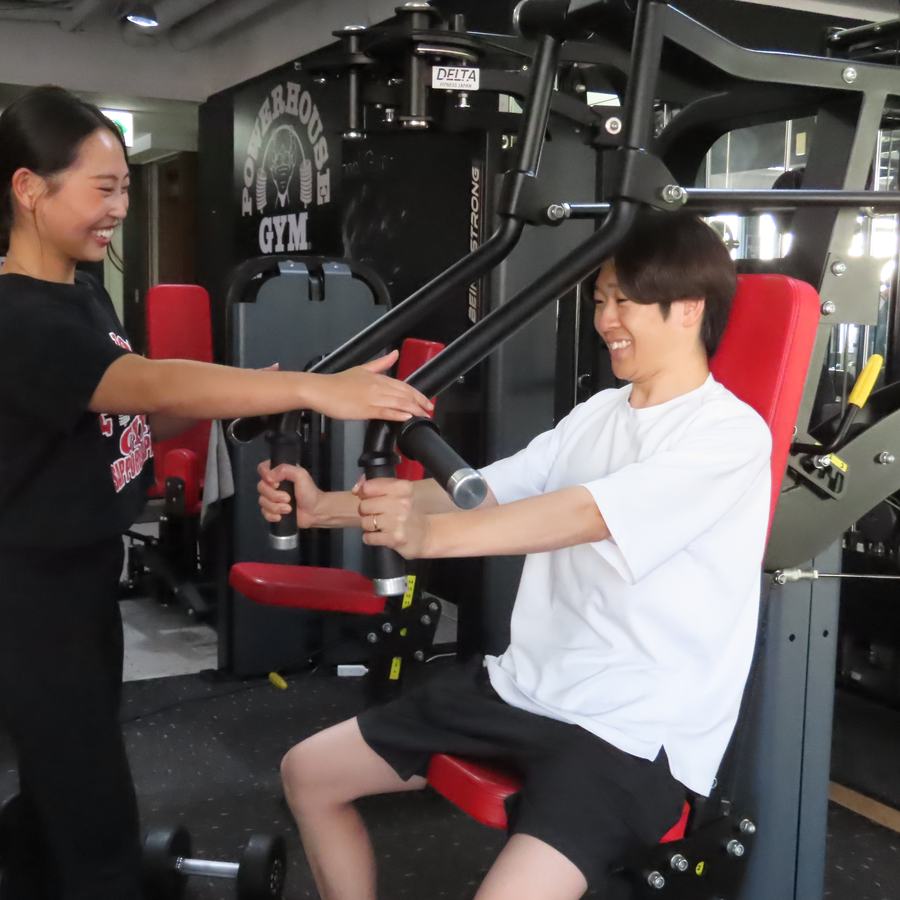 チェストプレスをする男性の横で、トレーナーの野村がマシンのアームに手を添えている