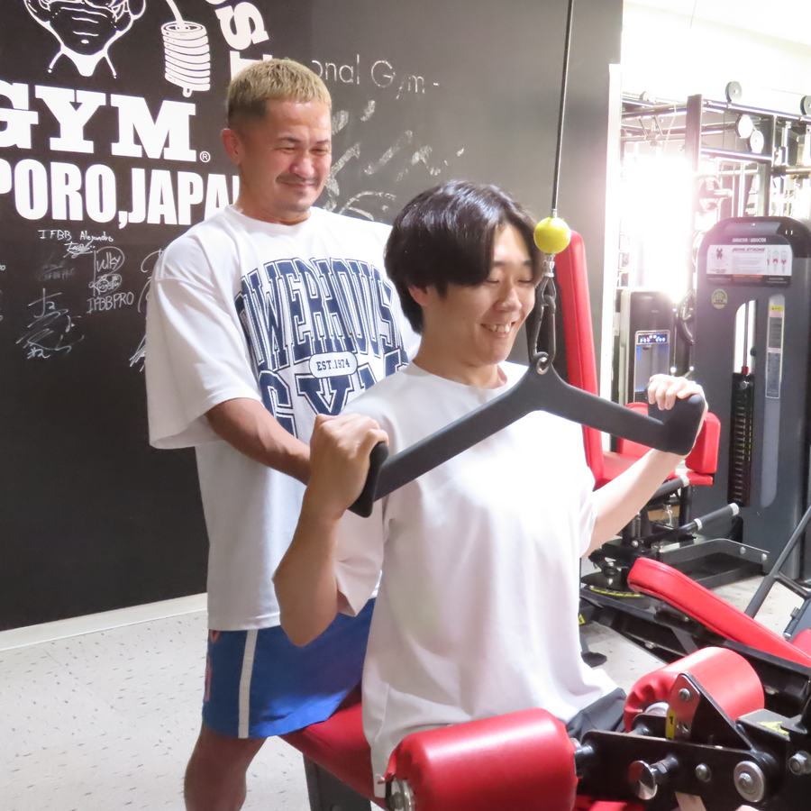 ラットプルダウンをする男性の後ろで、トレーナーの外崎が見守っている